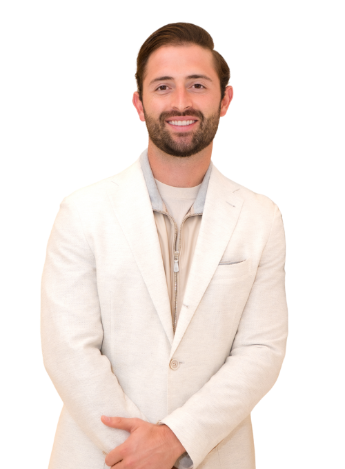 A smiling man with short brown hair and a beard, wearing a light beige blazer over a beige shirt, stands with his hands clasped in front of him against a white background.