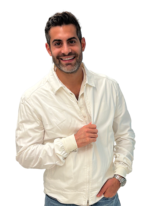 A man with short dark hair and a beard smiles at the camera. He is wearing a white jacket over a light shirt and jeans, with a watch on his wrist. He stands in a well-lit indoor setting.