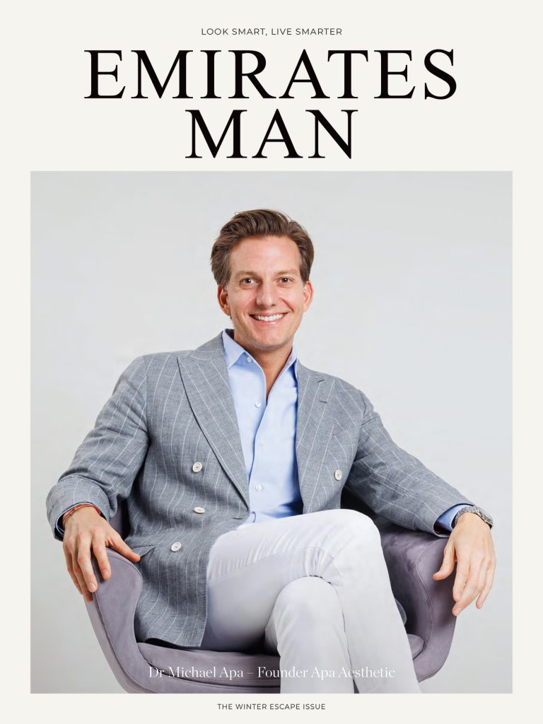 A man in a gray blazer and white pants sits confidently in a gray chair, smiling. The cover text reads Emirates Man with a feature on Dr. Michael Apa, Founder of Apa Aesthetic. The theme is The Winter Escape Issue.