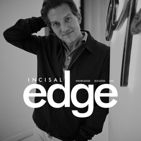 A man in a dark jacket leans against a wall, looking at the camera with a slight smile. His reflection is visible in the shiny surface next to him. The magazine cover features Incisal Edge in large white letters.