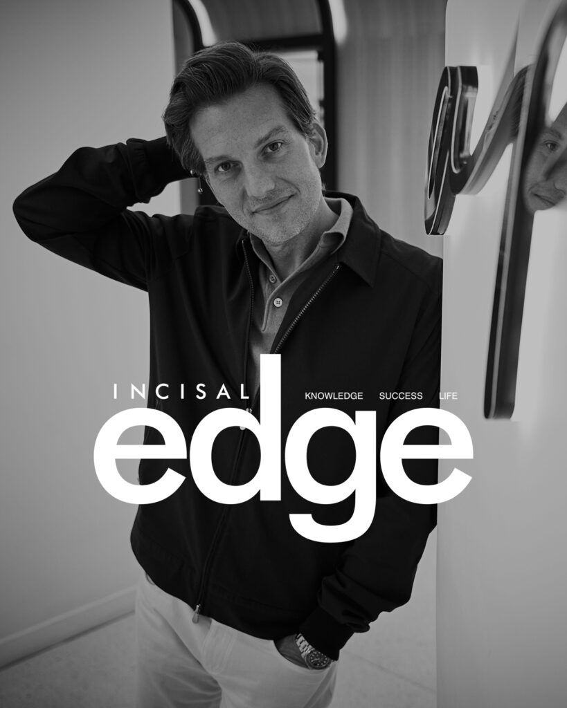 A man in a dark jacket leans against a wall, looking at the camera with a slight smile. His reflection is visible in the shiny surface next to him. The magazine cover features Incisal Edge in large white letters.