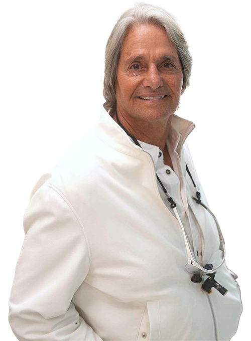 A person with grey hair smiles while wearing a white jacket and a white shirt. A pair of magnifying glasses hangs around their neck.