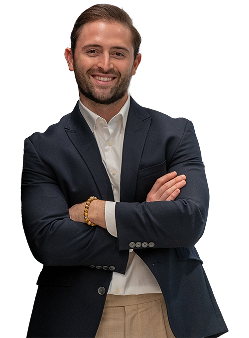 A smiling man with short hair and a beard is wearing a dark blazer over a white shirt. He is standing with his arms crossed and one hand is adorned with a beaded bracelet. The background is plain white.