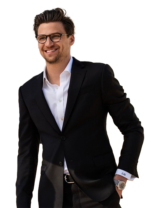 A smiling man wearing glasses and a black suit with a white shirt. He has his left hand in his pocket. His suit jacket is open, and he is standing against a plain background.