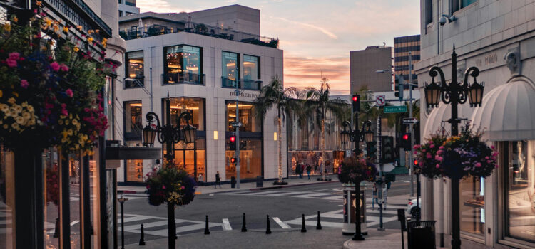 A quiet city street at sunset with elegant buildings and streetlights adorned with hanging flower baskets. Pedestrians stroll by upscale shops, and soft sunlight illuminates the scene, creating a warm, inviting atmosphere.