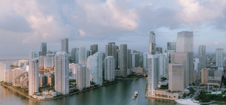 Aerial view of a cityscape with modern high-rise buildings surrounded by water. The skyline features a mix of rectangular and curved architectural designs. The sky is cloudy, and a boat travels through the waterway.