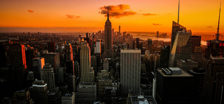 A breathtaking cityscape of New York at sunset, with skyscrapers silhouetted against a vibrant orange sky. The iconic Empire State Building stands prominently in the center, surrounded by other tall buildings and a distant view of the horizon.