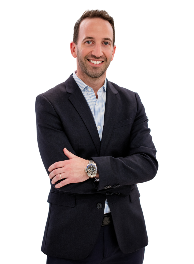 A man in a dark suit and a light blue shirt stands with his arms crossed, smiling. He wears a watch on his left wrist. The background is plain white.