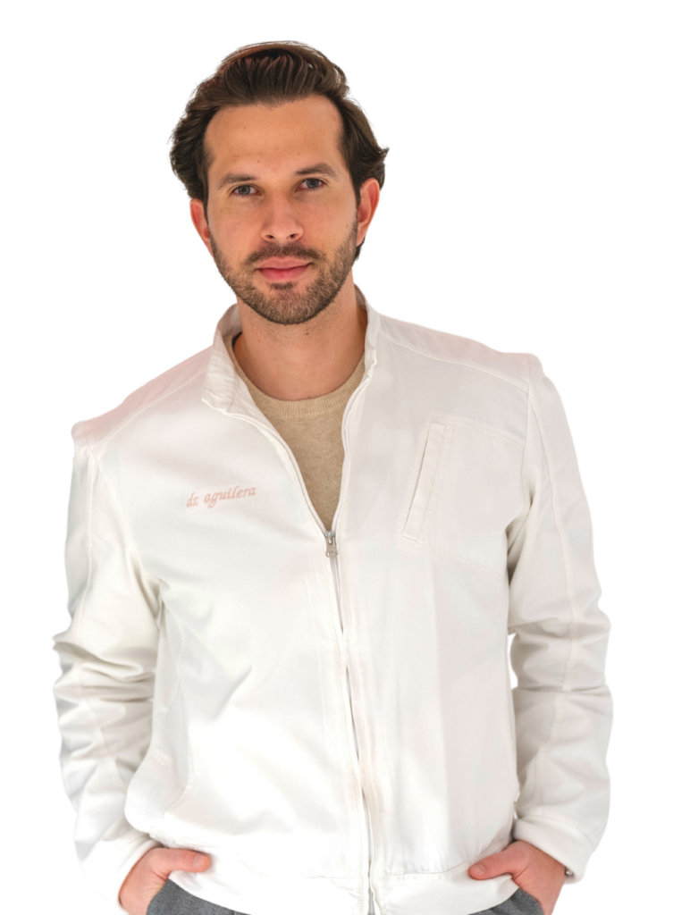 A person with short dark hair and a trimmed beard is standing against a white background, wearing a light-colored jacket with hands in pockets and looking forward.