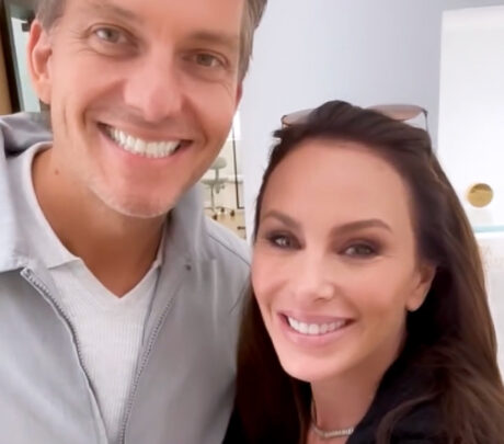 A man and a woman are smiling at the camera. The man is wearing a light jacket over a white shirt, and the woman has long hair and is wearing a necklace. They appear to be indoors.