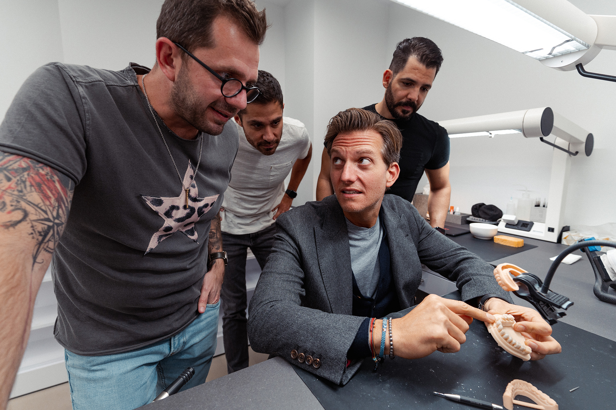 Four men are gathered around a seated man, who is holding and examining a dental mold. They appear to be discussing or learning about dental techniques in a modern, well-lit workspace.