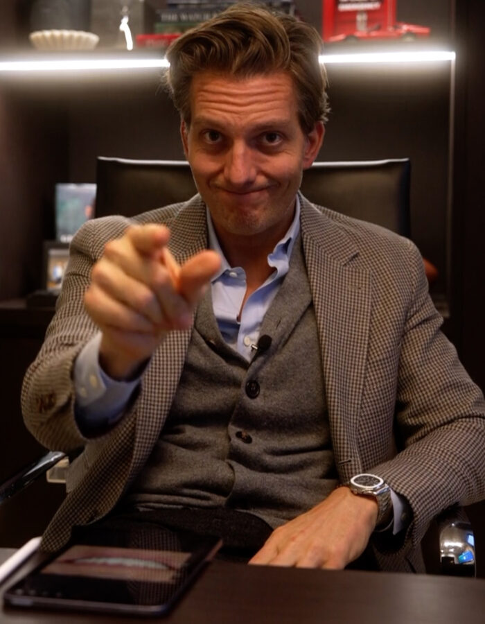 A man in a suit sits at an office desk, smiling and pointing at the camera. Behind him are shelves with various decorative items.