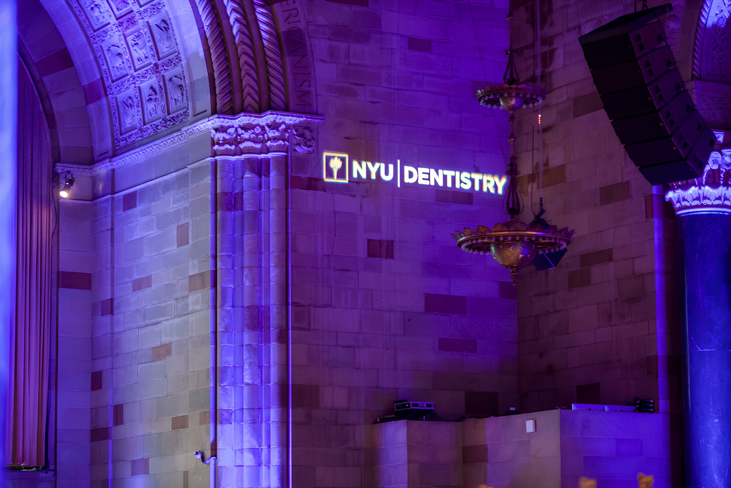 A grand interior with ornate architecture and purple lighting. A sign on the wall reads NYU Dentistry, illuminated in white and yellow. The space features archways and decorative chandeliers.