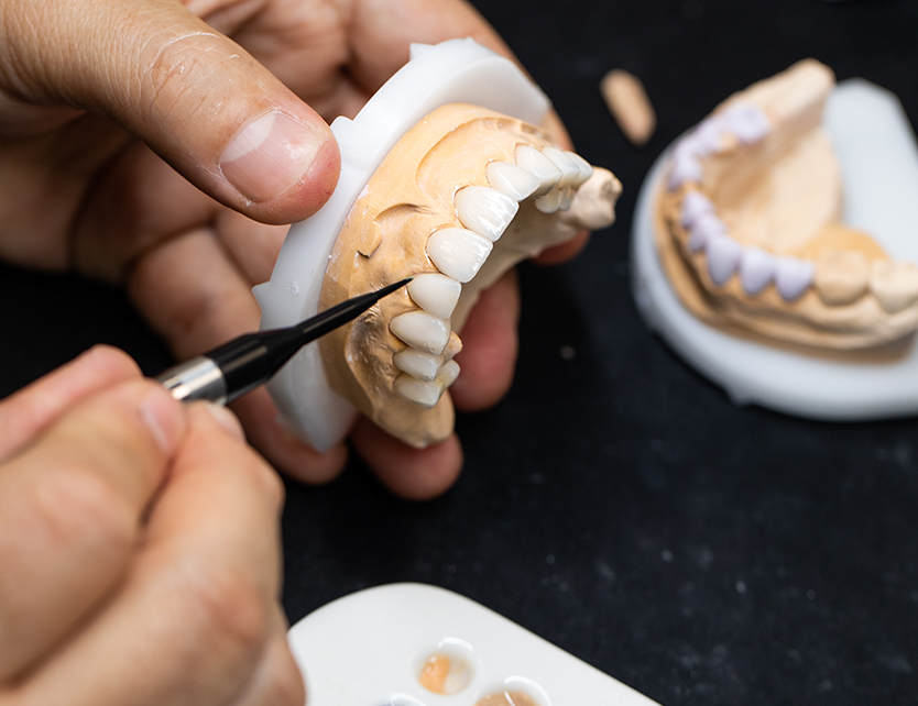 A person uses a tool to carefully paint color onto a set of dental prosthetic teeth on a model. Another model with different shades is visible in the background.