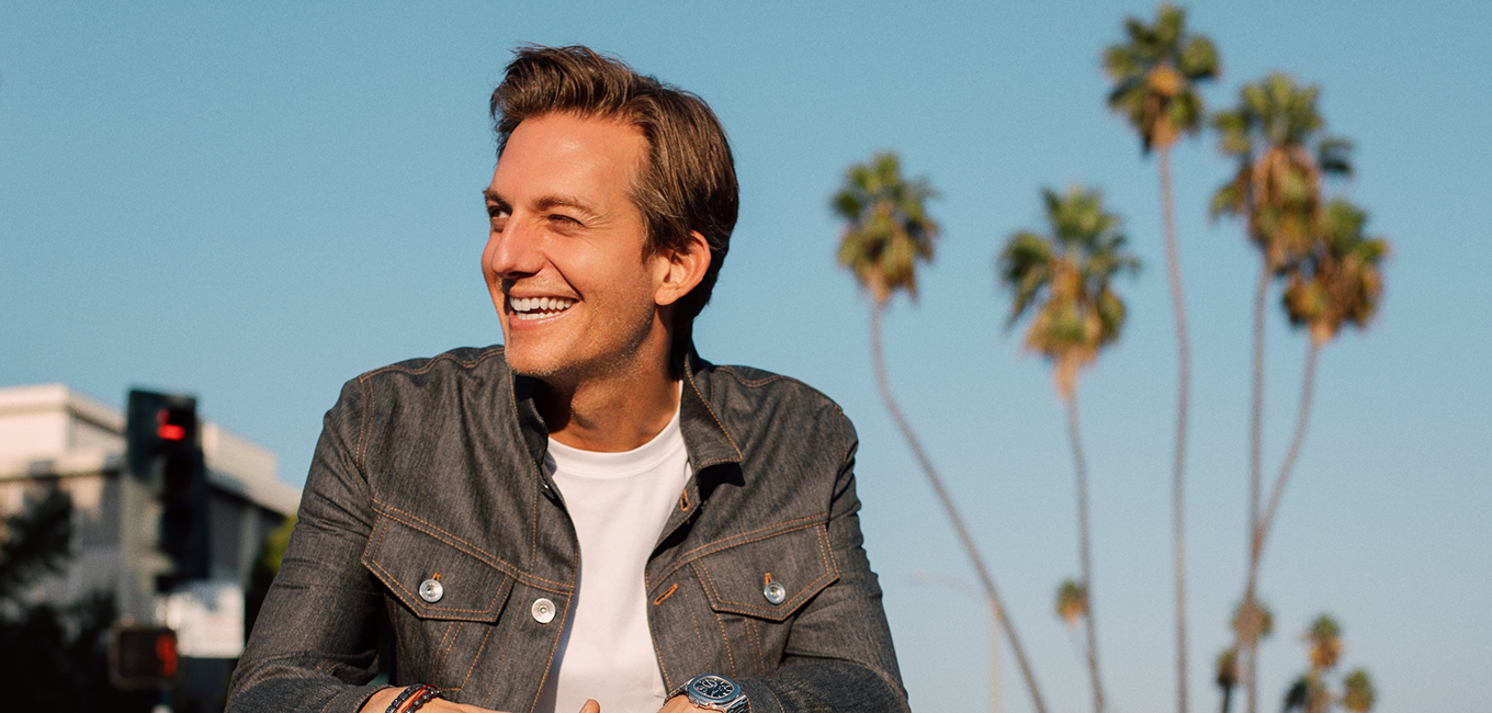 A smiling man wearing a denim jacket and white shirt stands outdoors under a clear blue sky. Tall palm trees are visible in the background, and a traffic light is on the left.