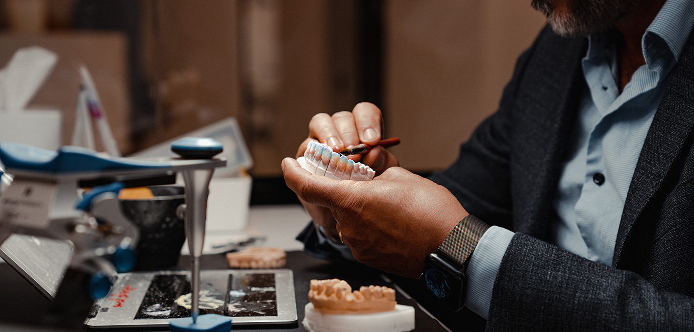 A person carefully paints a dental prosthetic model at a workstation. The scene includes various dental tools and models, with the individuals hands focused on detailing the prosthetic.