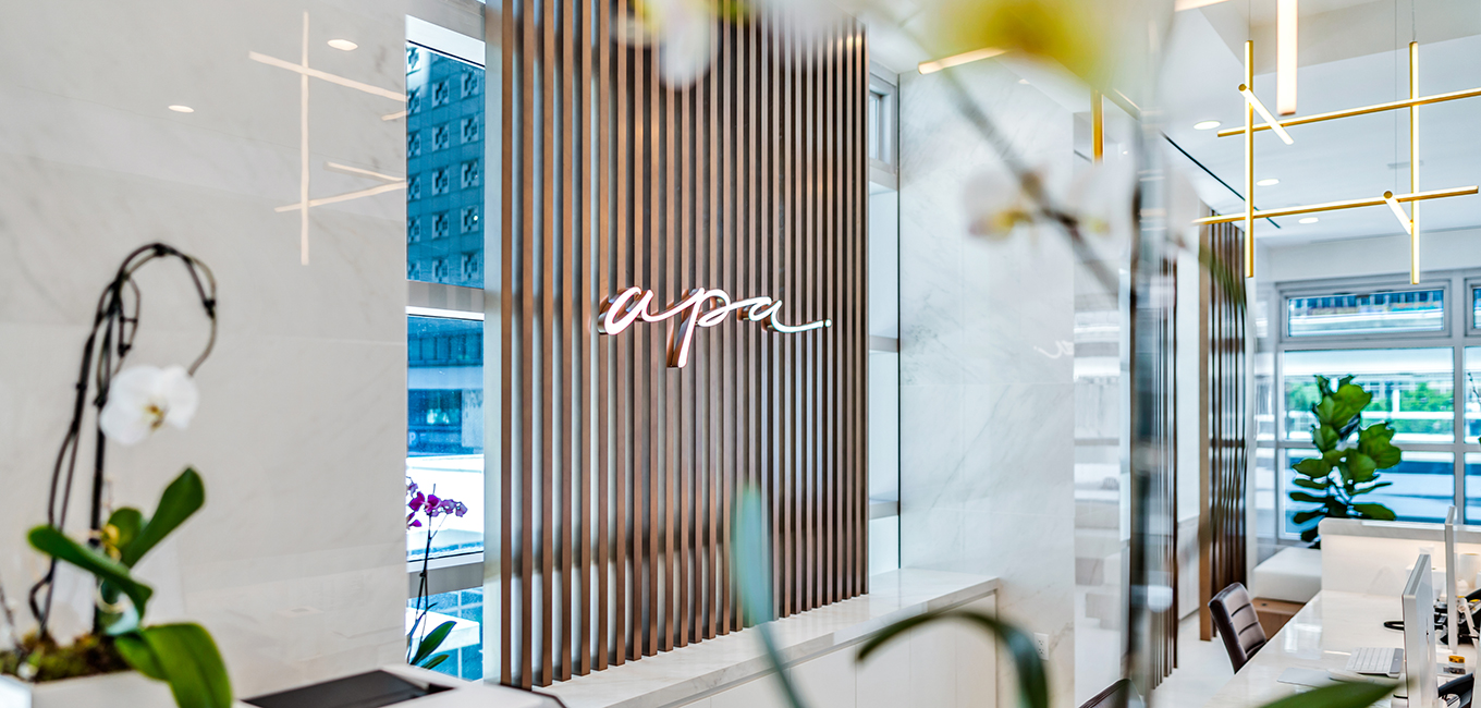 Bright, modern office space featuring a wall with vertical wooden slats and a pink neon sign that reads apa. The interior has marble finishes, large windows, and potted plants.