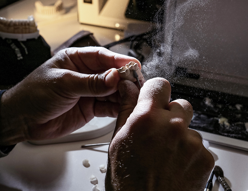 Hands working on a dental model in a lab, with dust particles illuminated by light. Tools and dental molds are visible on the table in the background.