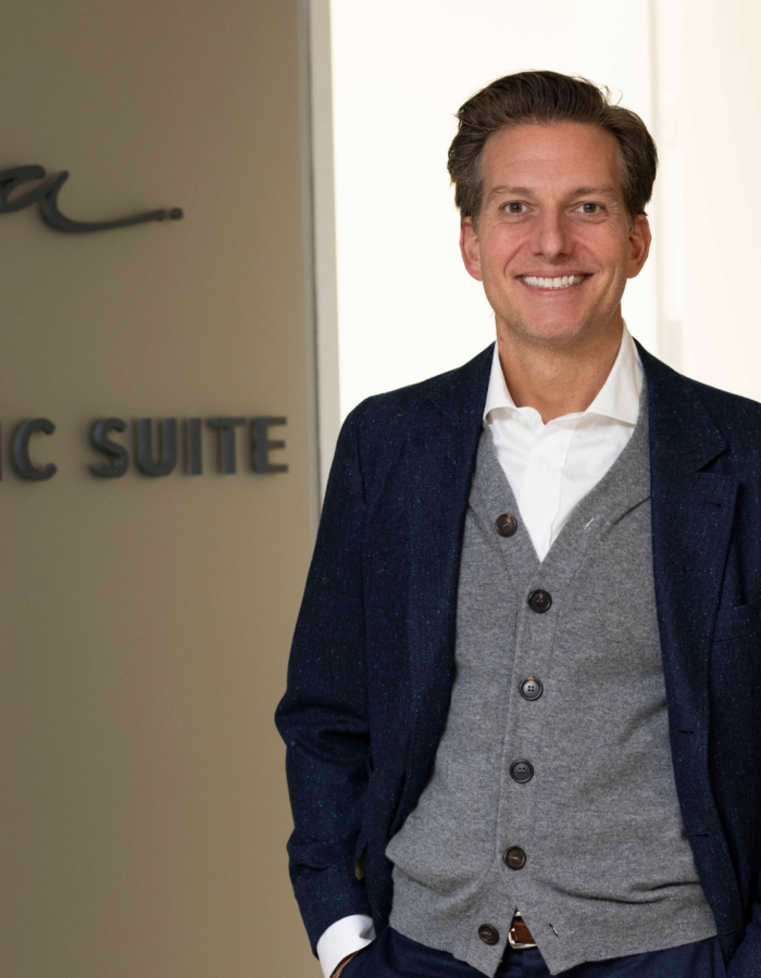 Smiling man in a gray cardigan and dark blazer stands indoors near a wall with partial lettering visible.