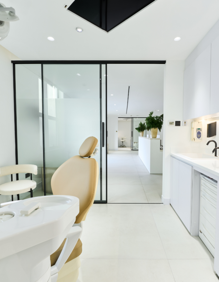 Modern dental office with a beige dental chair facing a frosted glass sliding door. The room features white cabinets, a black ceiling panel, and a view into a bright hallway with plants.