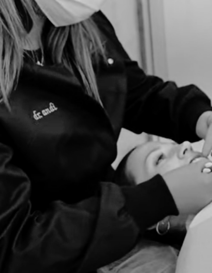 A dental professional wearing a mask and magnifying glasses examines a patients teeth. The patient is lying back in a dental chair, with their mouth open, receiving care. The scene is in a clinical setting.