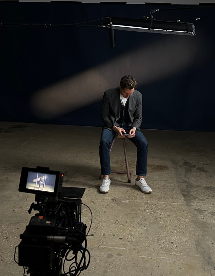 A man in a blazer and jeans sits on a stool under a spotlight, looking down at his phone. A boom microphone hangs above him, and a camera and headphones are visible in the foreground on a concrete floor.