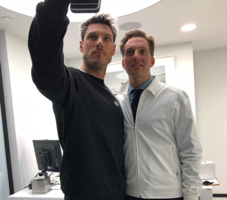 Two men stand side by side in a modern, well-lit office, posing for a selfie. One wears a black sweatshirt and holds the phone, while the other wears a white jacket and tie, both looking at the camera.