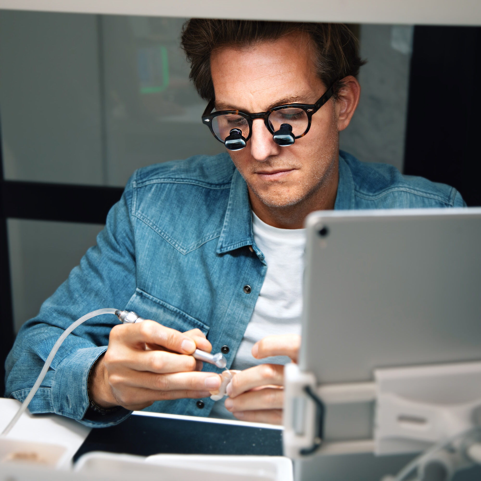 A person in a denim shirt is using a dental tool on a model while wearing magnifying glasses. They are focused on their work at a table with a tablet computer in a holder nearby.