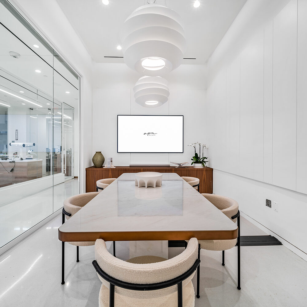 Modern conference room with a long marble table surrounded by beige chairs. Two large white pendant lights hang from the ceiling. A flat-screen TV is mounted on the back wall, and a glass partition reveals an office area.