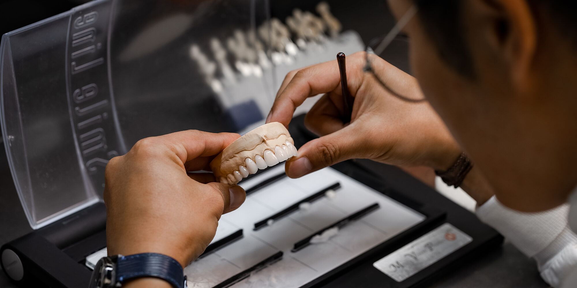 A person examines a dental model with a small tool, seated at a workstation. Various dental instruments are organized in front of them, and more dental models are visible in the background.