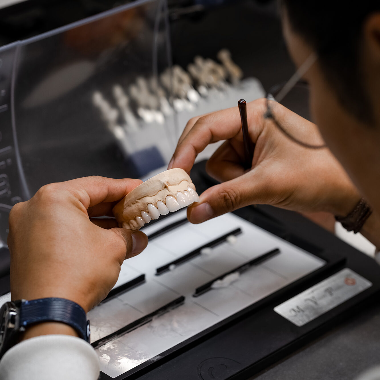 A person examines a dental model with a small tool, seated at a workstation. Various dental instruments are organized in front of them, and more dental models are visible in the background.