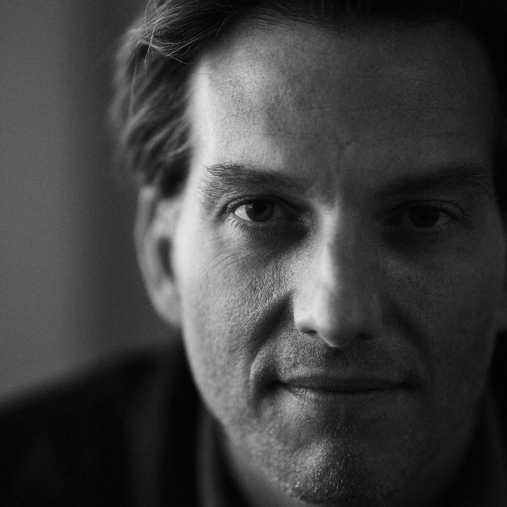 A black and white close-up portrait of a man with short hair, looking directly at the camera. The lighting highlights his facial features against a softly blurred background.