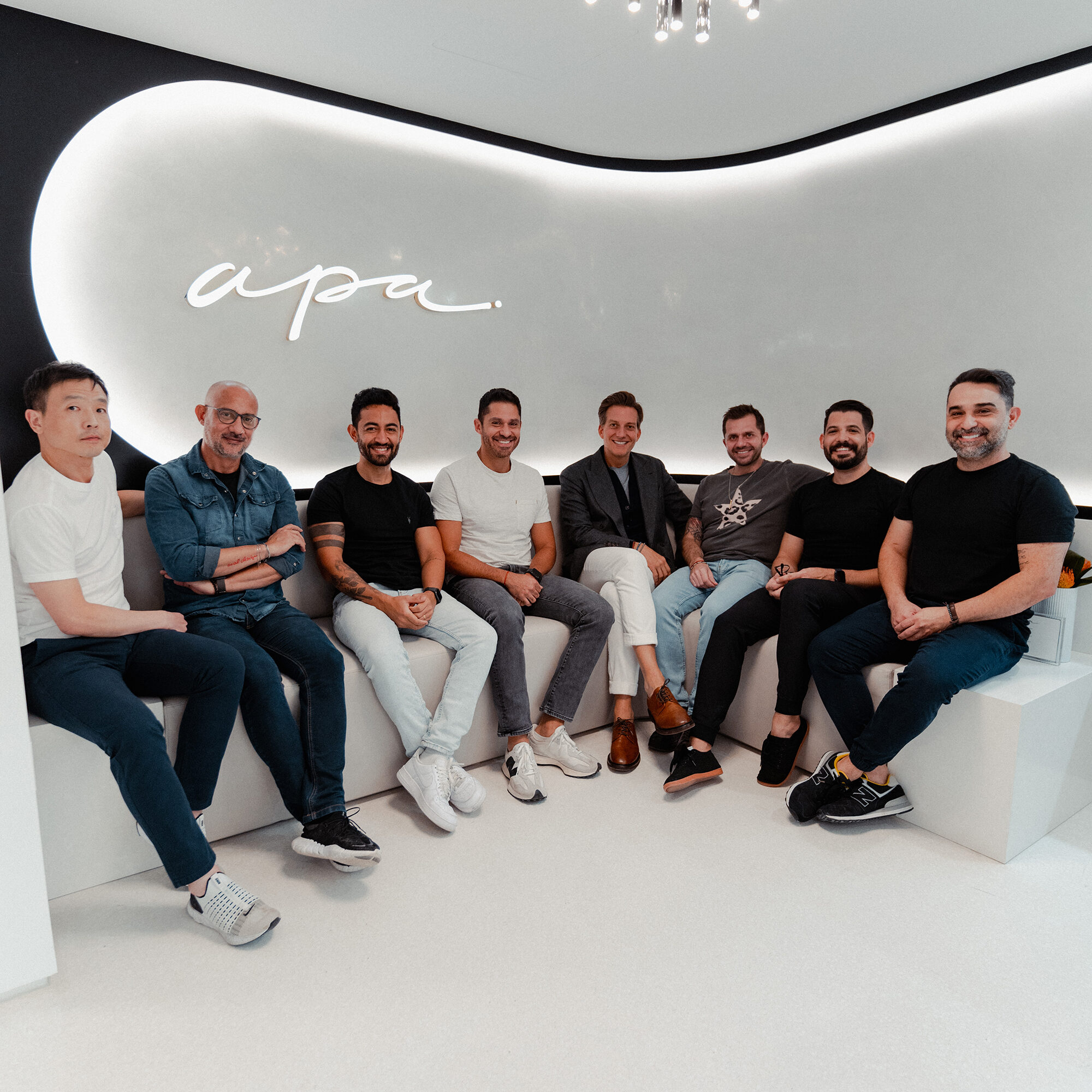 A group of eight men sitting together on white benches in a modern, well-lit room with a sleek design. The wall features a large, stylized apa logo. They are casually dressed and smiling at the camera.