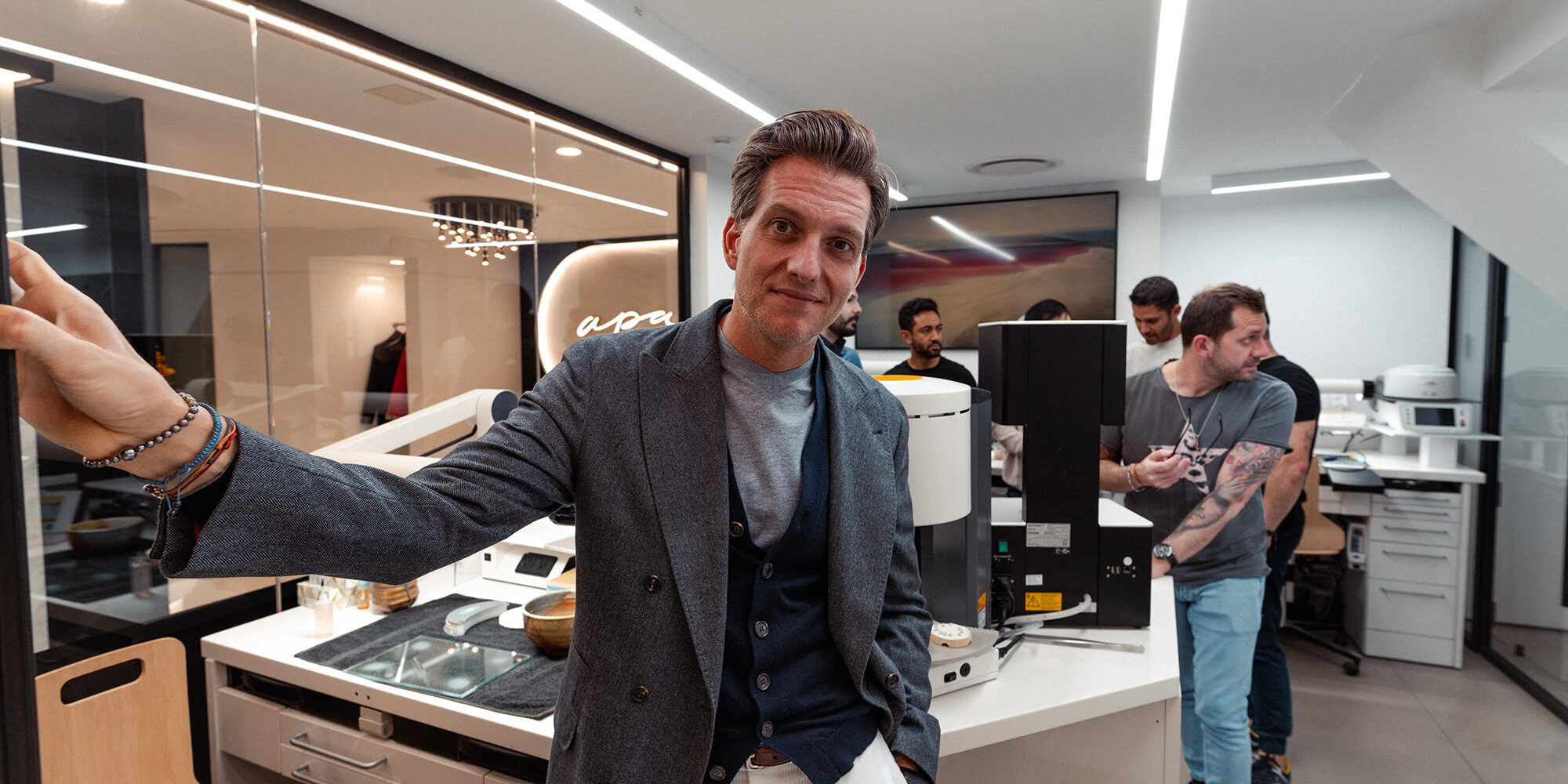 A man in a gray blazer and white pants leans casually against a desk in a modern office with several people working in the background, some using computers and equipment. The space is well-lit with sleek, contemporary design elements.