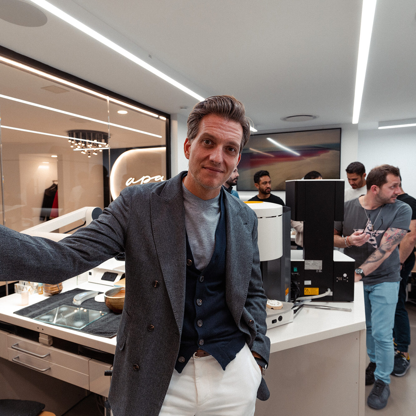 A man in a gray blazer and white pants leans casually against a desk in a modern office with several people working in the background, some using computers and equipment. The space is well-lit with sleek, contemporary design elements.